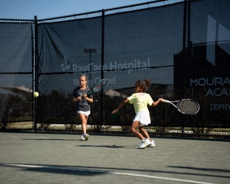 Two young girls playing tennis on an outdoor court at Mouratoglou Academy Zephyrhills on a sunny day.