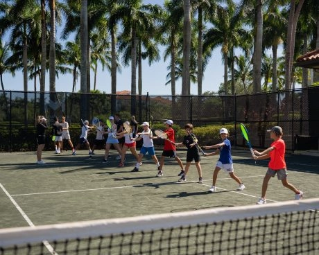 Group tennis lesson for kids practicing forehand drills on an outdoor clay court with palm trees.