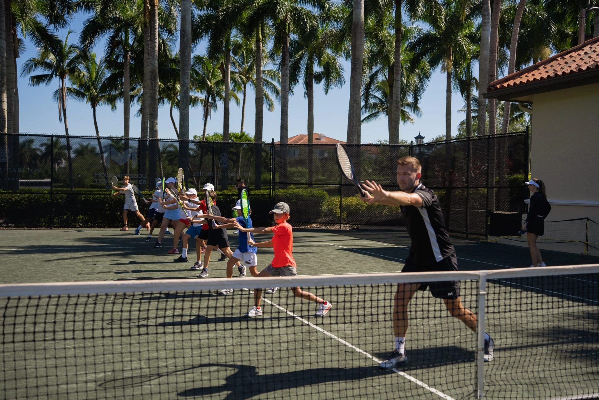 Group tennis lesson for kids practicing forehand drills on an outdoor clay court with palm trees