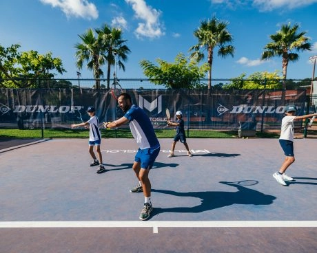 Tennis coach leading a group drill with young players on an outdoor court under palm trees