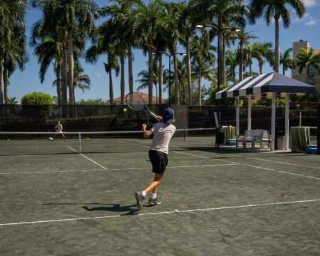 Young tennis player hitting a forehand on an outdoor clay court surrounded by palm trees