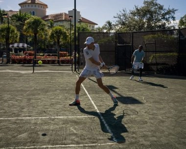 Tennis players practicing on an outdoor clay court surrounded by palm trees in sunny weather.