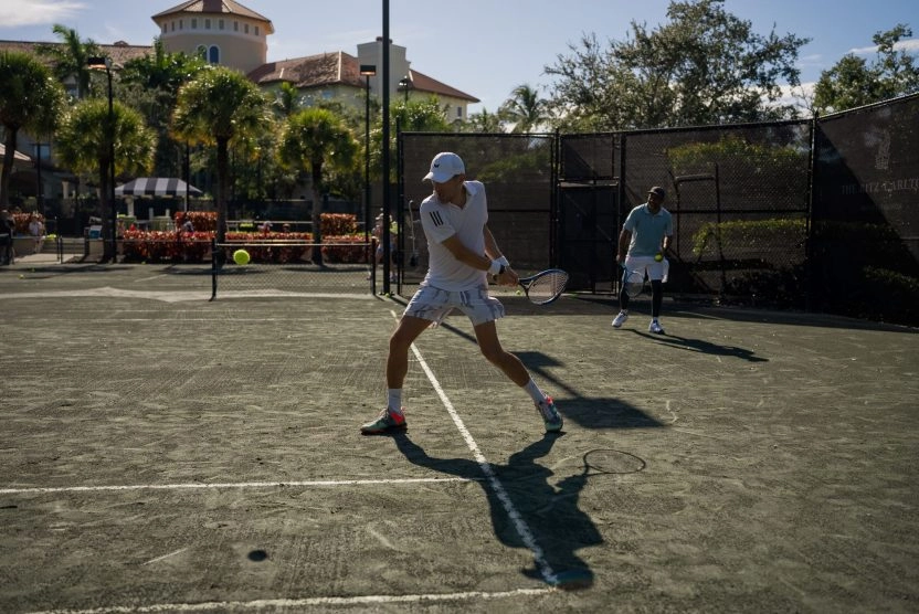 Tennis players practicing on an outdoor clay court surrounded by palm trees in sunny weather.
