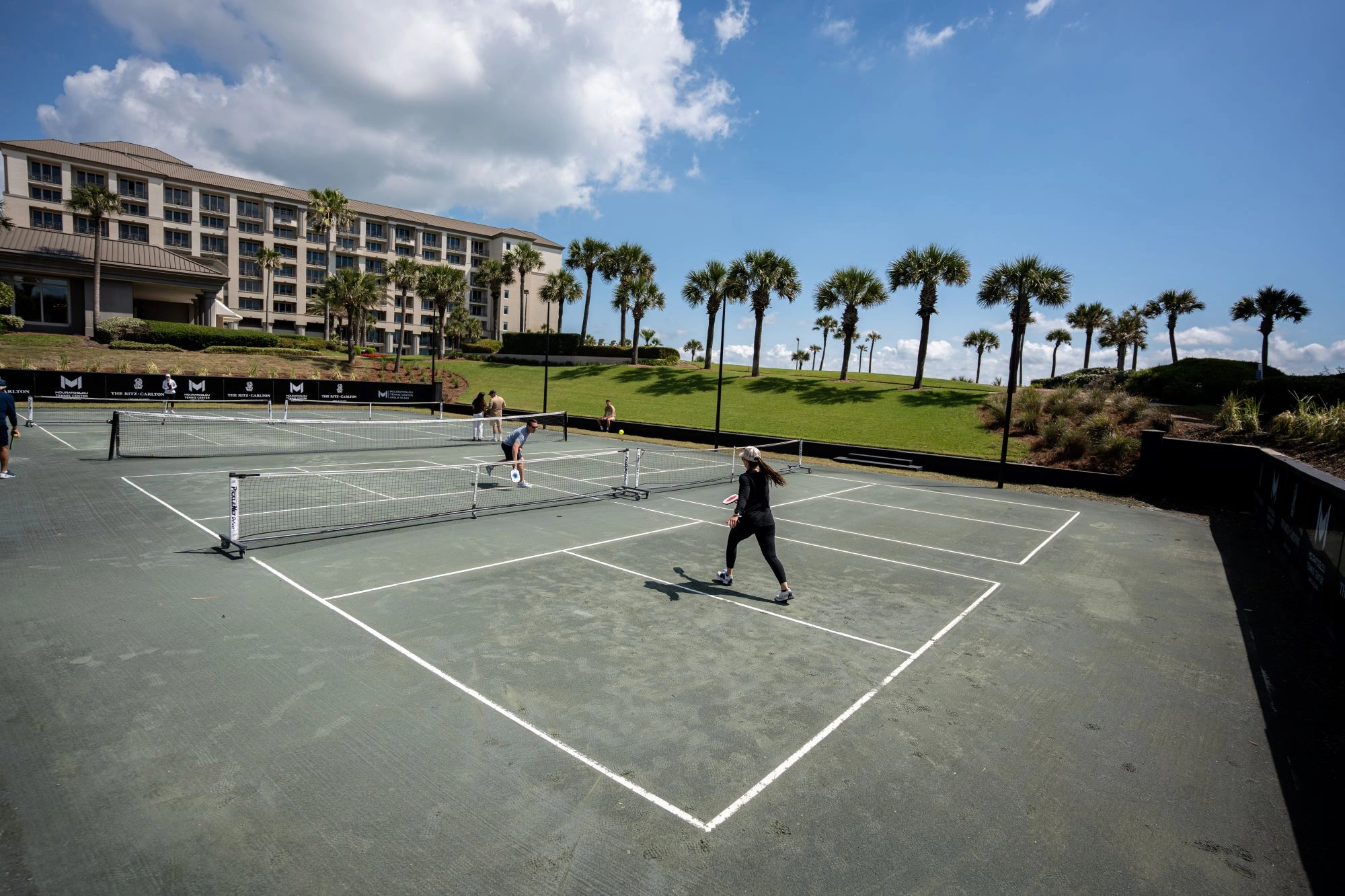 Players practice on green Har-Tru courts at Mouratoglou Tennis Center Amelia Island, with palm trees and resort buildings behind them.