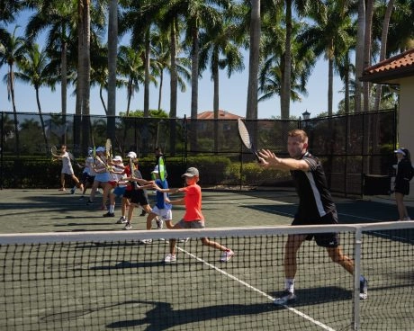 Group tennis lesson for kids practicing forehand drills on an outdoor clay court with palm trees