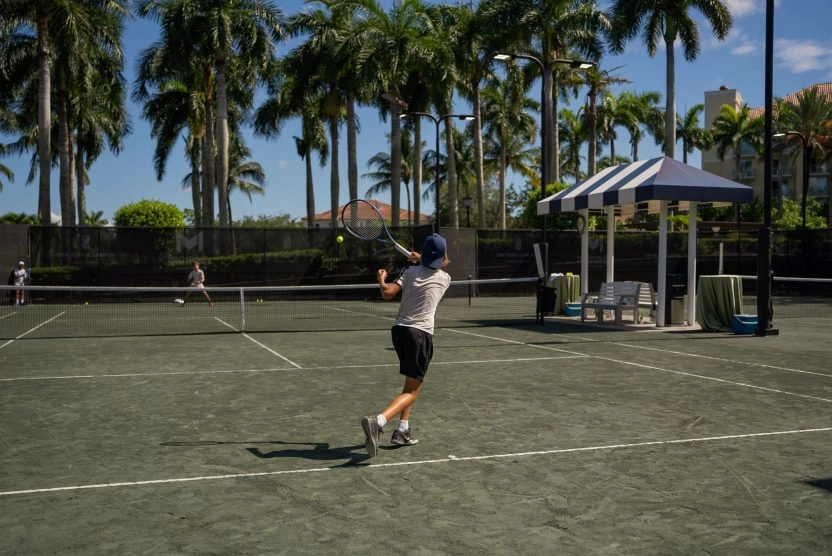 Young tennis player hitting a forehand on an outdoor clay court surrounded by palm trees