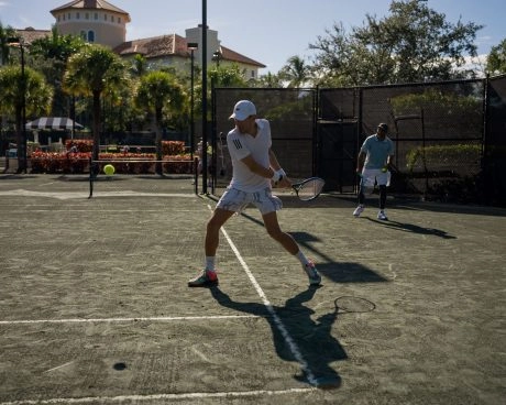 Tennis players practicing on an outdoor clay court surrounded by palm trees in sunny weather