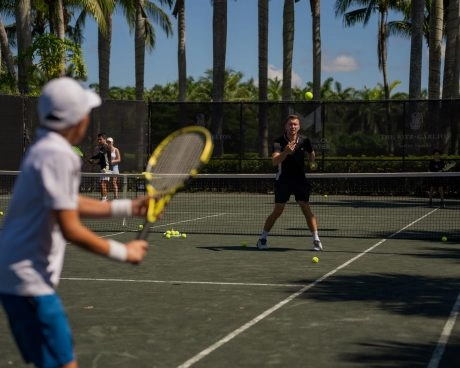 Tennis coach training a young player on an outdoor clay court surrounded by palm trees