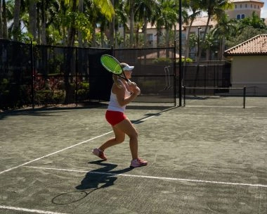 Female tennis player hitting a backhand shot on an outdoor clay court surrounded by palm trees.