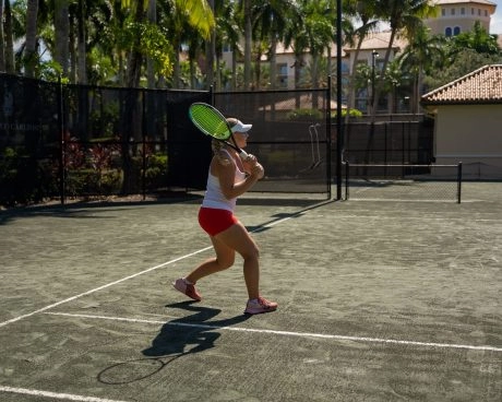 Female tennis player hitting a backhand shot on an outdoor clay court surrounded by palm trees.