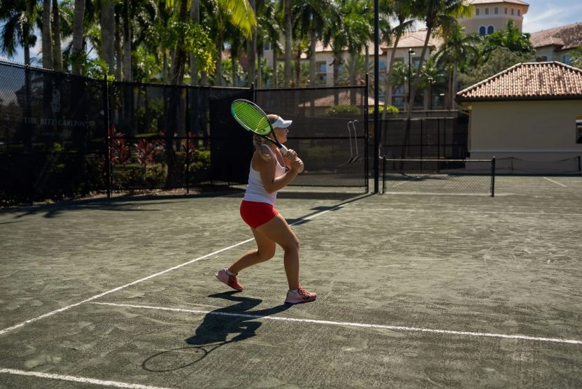 Female tennis player hitting a backhand shot on an outdoor clay court surrounded by palm trees.
