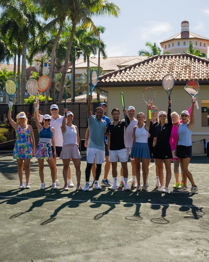 Group of adult tennis players posing with raised rackets on an outdoor court at a sunny resort