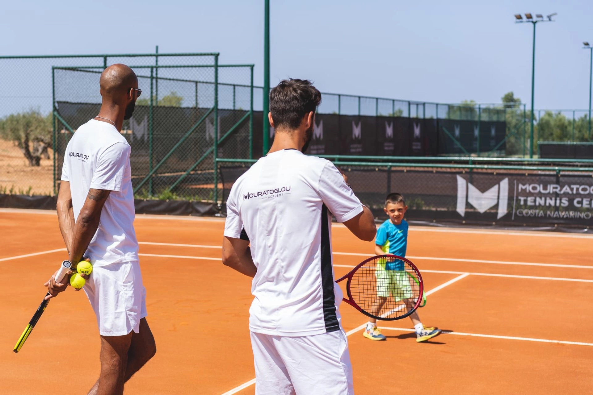 Tennis coaches guiding a young child during training on a clay court.