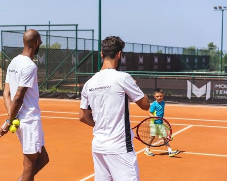 Tennis coaches guiding a young child during training on a clay court.