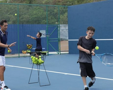 Tennis coach feeding balls to a young player practicing a forehand on a blue court