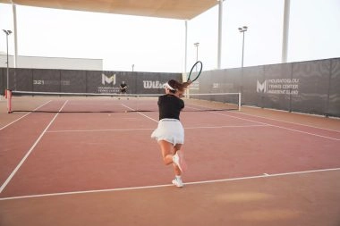 Female tennis player hitting a forehand on the courts of Mouratoglou Tennis Center Hudayriyat Island in Abu Dhabi, surrounded by sea breeze and sunlight.