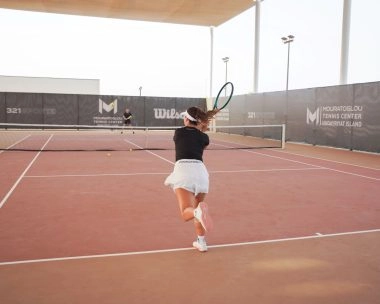 Female tennis player hitting a forehand on the courts of Mouratoglou Tennis Center Hudayriyat Island in Abu Dhabi, surrounded by sea breeze and sunlight.
