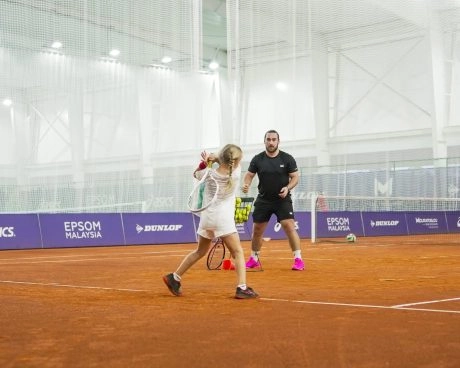 Young tennis player practicing a backhand on an indoor clay court with a coach.