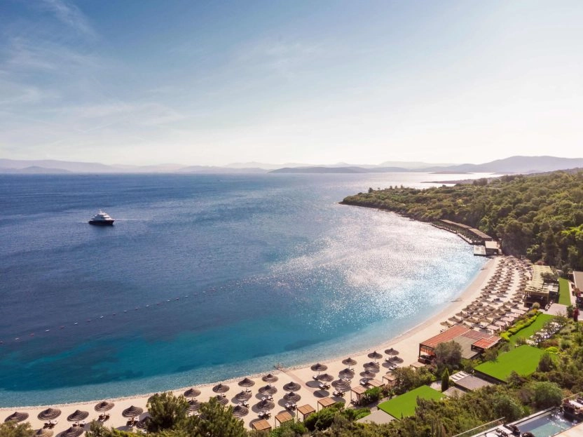 Seaside view of the Bodrum coastline with a clear blue sea, beach umbrellas, and lush greenery.