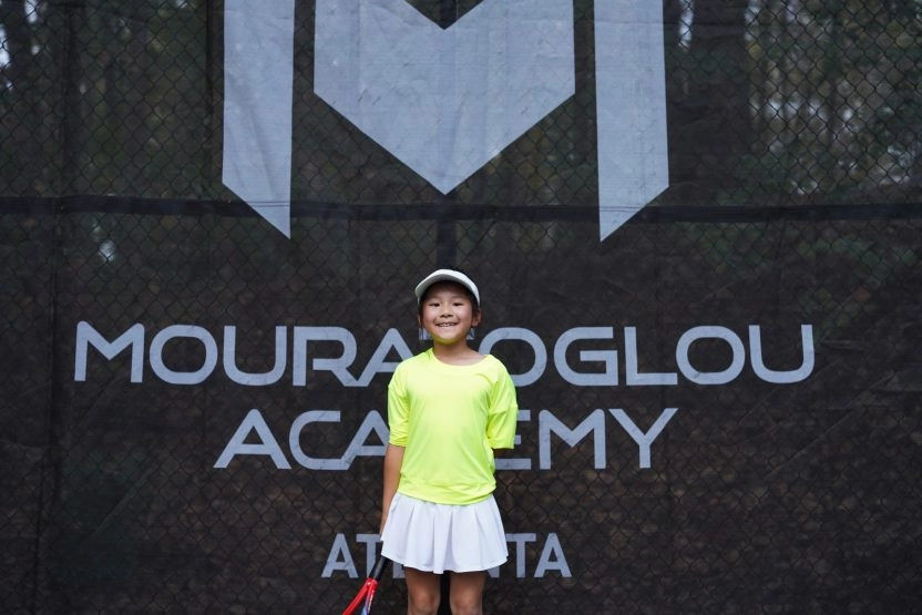 Smiling young girl standing in front of Mouratoglou windbreakers at Mouratoglou Academy Atlanta