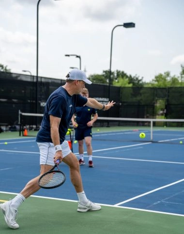 A man preparing a forehand during a private tennis lesson with a coach visible behind him at Mouratoglou Tennis Center Horseshoe Bay.