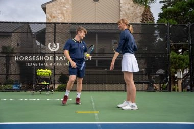 Two adults talking on a hard tennis court at Mouratoglou Tennis Center Horseshoe Bay.