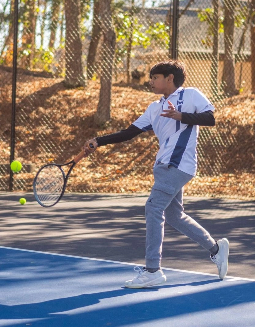 A young boy hitting a forehand shot while playing tennis on an outdoor court surrounded by trees.