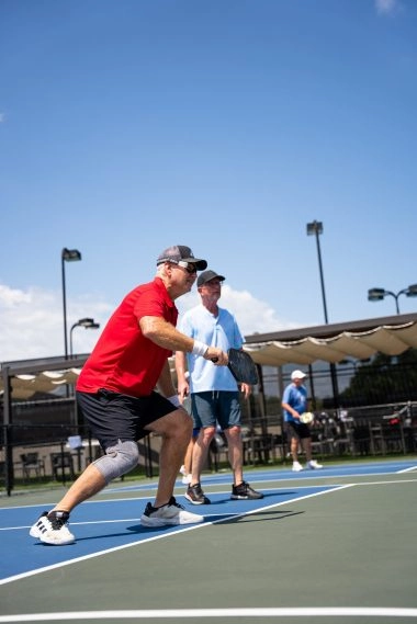 Deux adultes jouent un match de double sur un court de pickleball extérieur au Mouratoglou Tennis Center à Horseshoe Bay, sous le soleil.