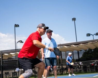 Deux adultes jouent un match de double sur un court de pickleball extérieur au Mouratoglou Tennis Center à Horseshoe Bay, sous le soleil.