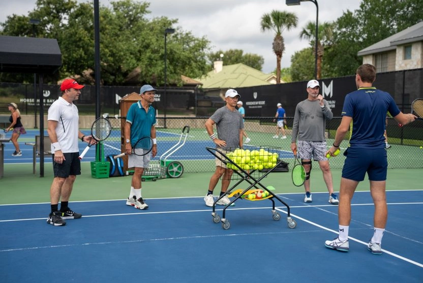 Tennis coach instructing a group of adult players on an outdoor court at Mouratoglou Tennis Center Horseshoe Bay with a cart of tennis balls.