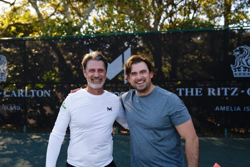 Patrick Mouratoglou posing with a player in front of Mouratoglou-branded windbreakers at Amelia Island.