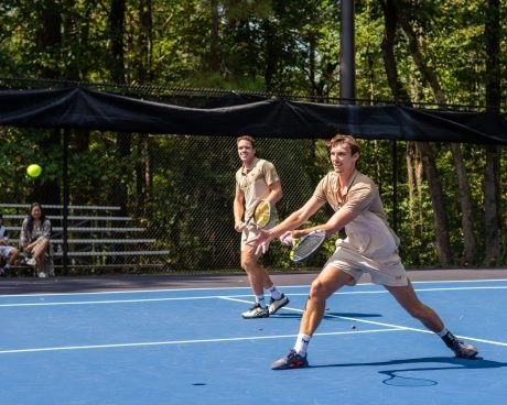 Two adult players competing in a doubles tennis match on an outdoor hard court at Mouratoglou Academy Atlanta with spectators in the background.