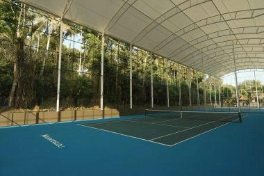 Semi-indoor blue hard tennis court at Mouratoglou Tennis Center Bali under a covered roof with surrounding tropical trees.