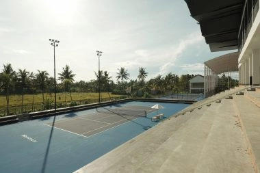 Outdoor hard tennis court at Mouratoglou Tennis Center Bali with spectator stands, umbrella seating, and tropical palm trees in the background.