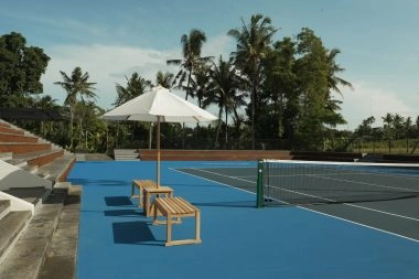 Outdoor hard tennis court at Mouratoglou Tennis Center Bali with spectator steps, benches under umbrella, and tropical palms.