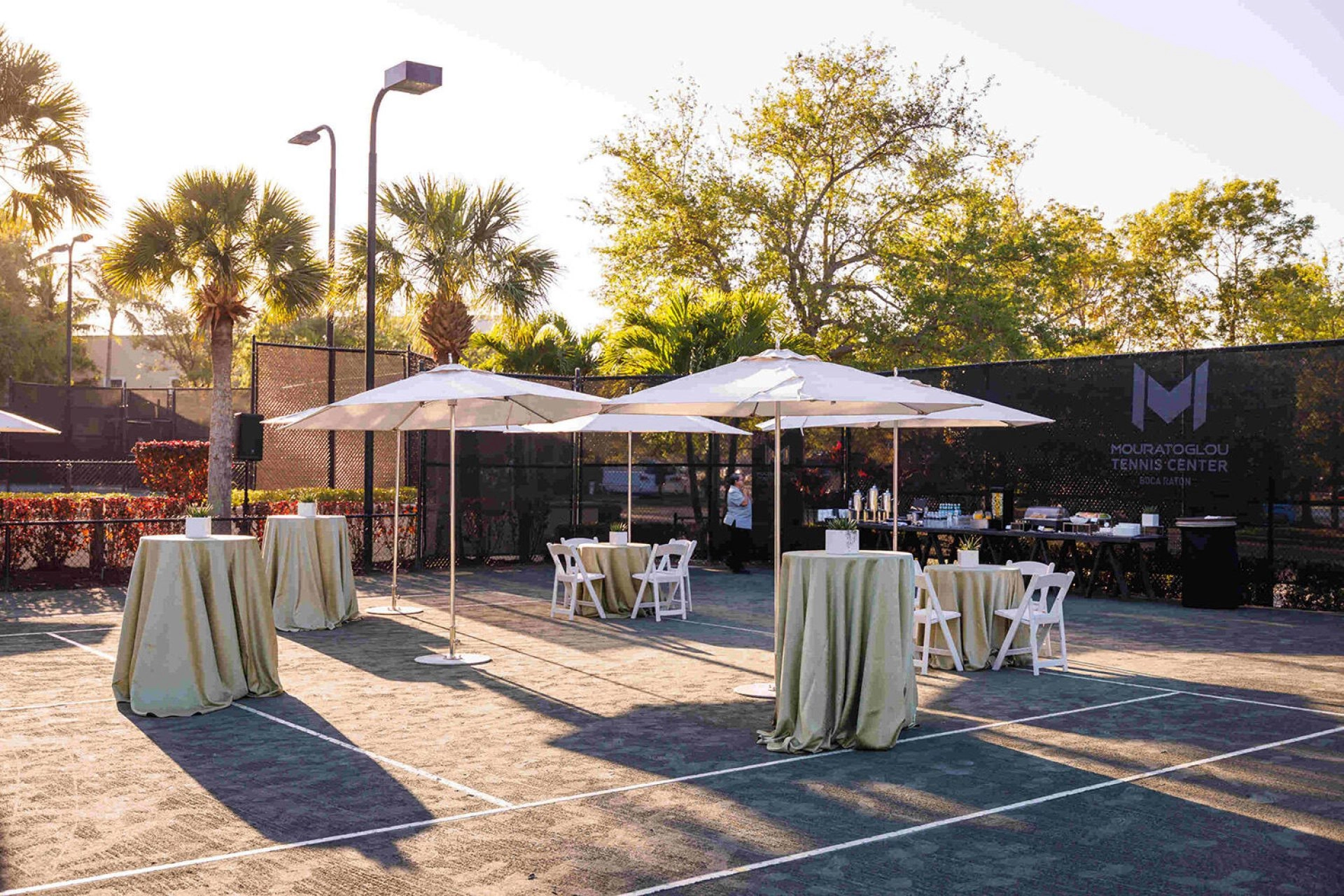Outdoor event reception setup on the courts at Mouratoglou Tennis Center Boca Raton, Florida, featuring hospitality stations and guest seating areas.