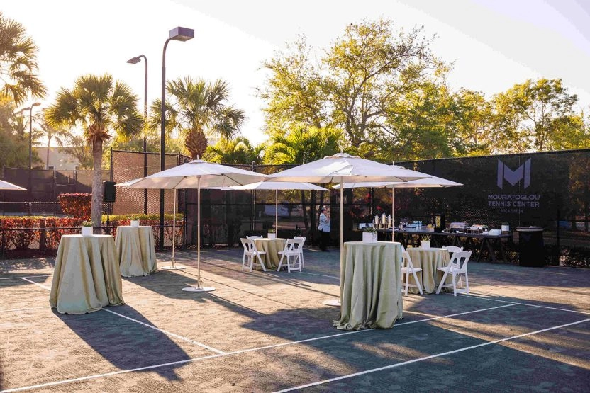 Outdoor event reception setup on the courts at Mouratoglou Tennis Center Boca Raton, Florida, featuring hospitality stations and guest seating areas.