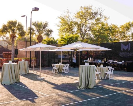 Outdoor event reception setup on the courts at Mouratoglou Tennis Center Naples, Florida, featuring hospitality stations and guest seating areas.