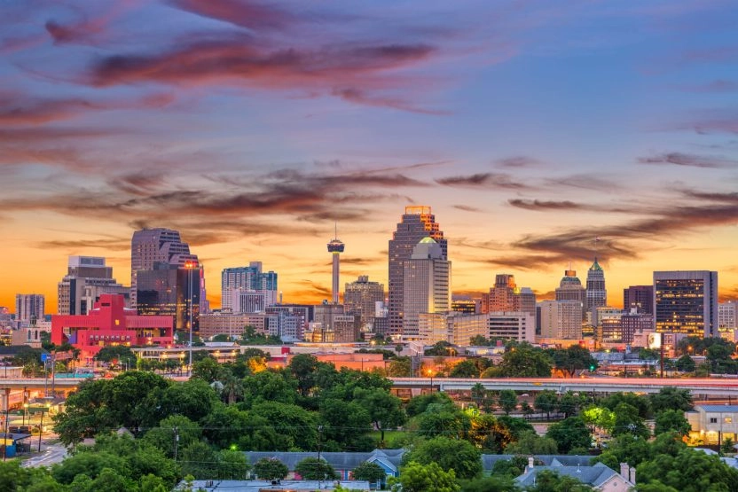 Downtown San Antonio skyline at sunset with high-rise buildings, green trees, and colorful clouds in the evening sky.