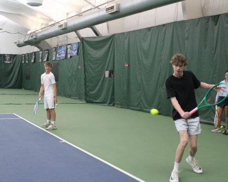 Tennis players practicing forehands on an indoor court at Mouratoglou Tennis Center Westchester with a coach and ball basket nearby.