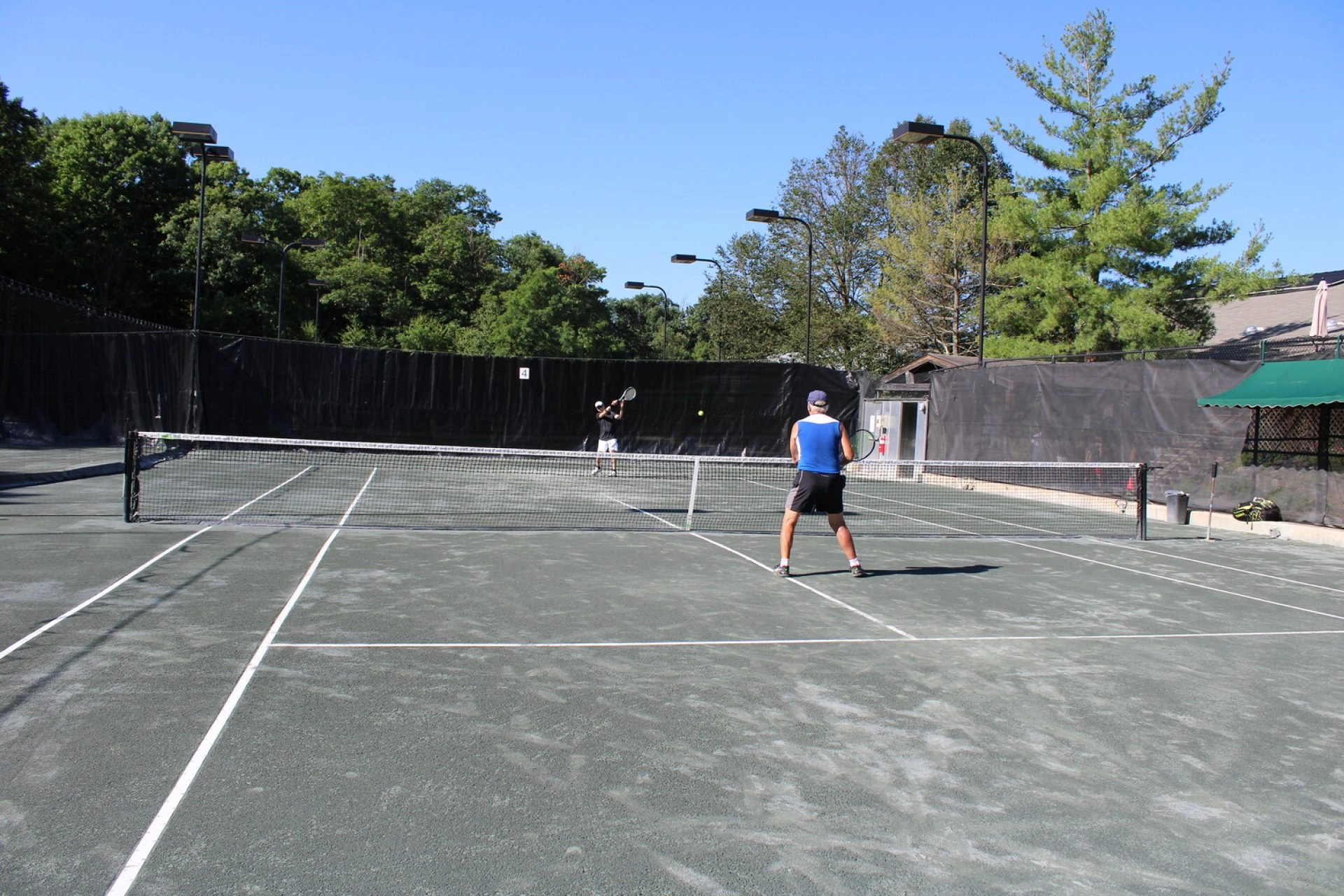 Two players rallying on an outdoor tennis court at Mouratoglou Tennis Center New York with trees and fencing in the background.