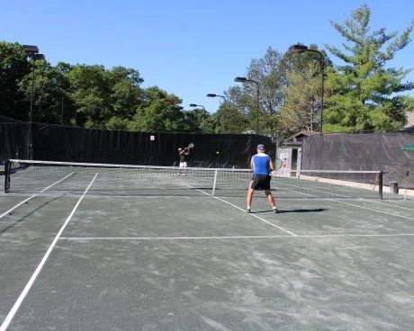 Two players rallying on an outdoor tennis court at Mouratoglou Tennis Center New York with trees and fencing in the background.