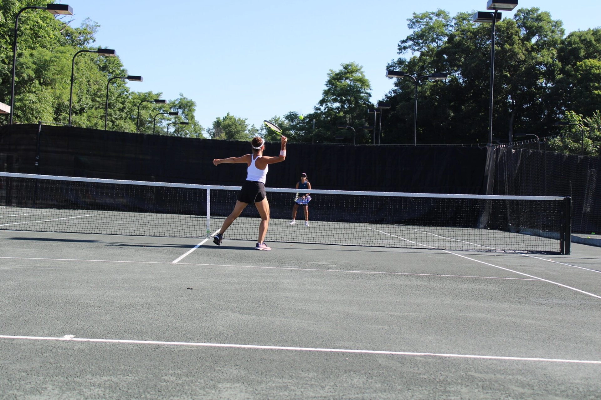Two players rallying on an outdoor tennis court at Mouratoglou Tennis Center Westchester with trees and fencing behind them.