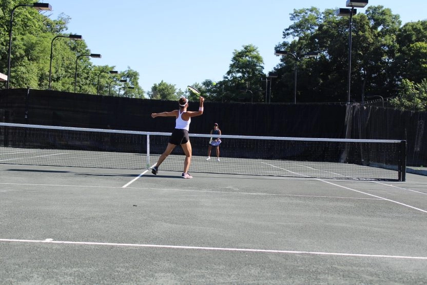 Two players rallying on an outdoor tennis court at Mouratoglou Tennis Center Westchester with trees and fencing behind them.