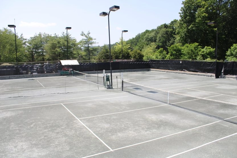Empty outdoor tennis courts at Mouratoglou Tennis Center Westchester with fencing, court lights, and trees in the background.