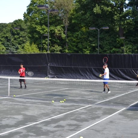 Tennis players practicing on an outdoor court at Mouratoglou Tennis Center Westchester with trees and fencing in the background.