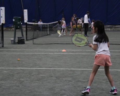 Young tennis player holding a racket on an indoor court at Mouratoglou Tennis Center Westchester with kids training in the background.