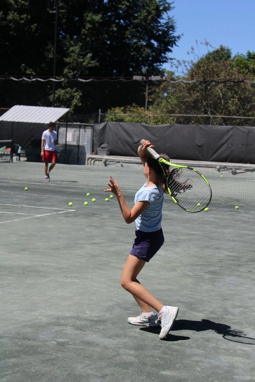 Young tennis player serving on an outdoor court at Mouratoglou Tennis Center Westchester with tennis balls and trees in the background.